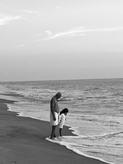 A child holding the hand of her father, feeling the sand under her soft feet for the first time, her eyes wide with wonder.